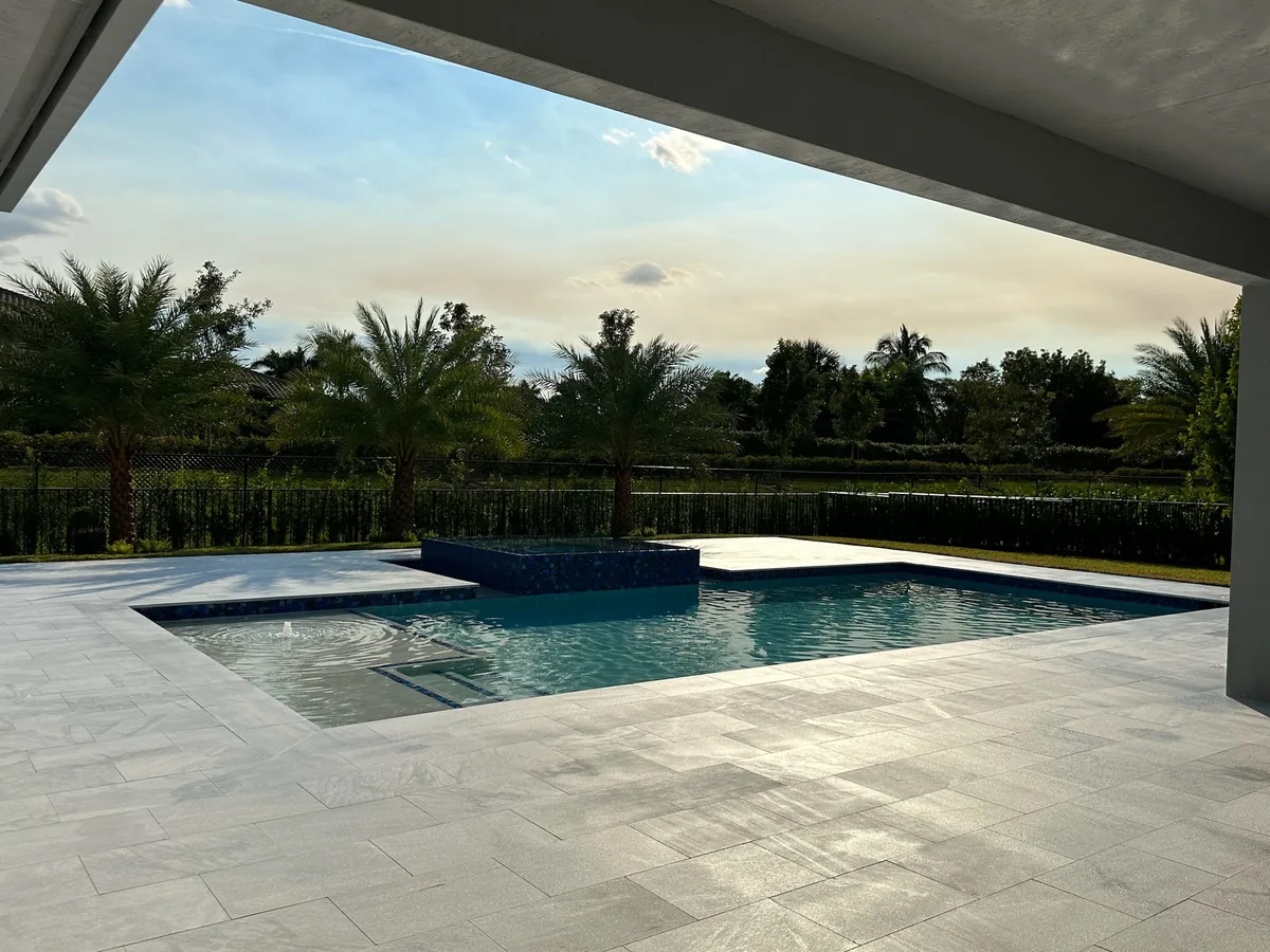 View from a covered patio over a pool with a raised spa and light paver deck at dusk