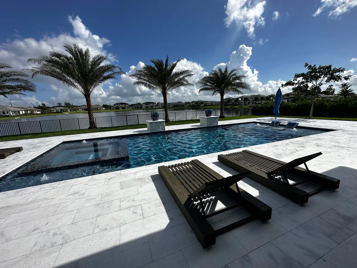 Wide view of a pool and spa with dark loungers, palms and a lake on a sunny day